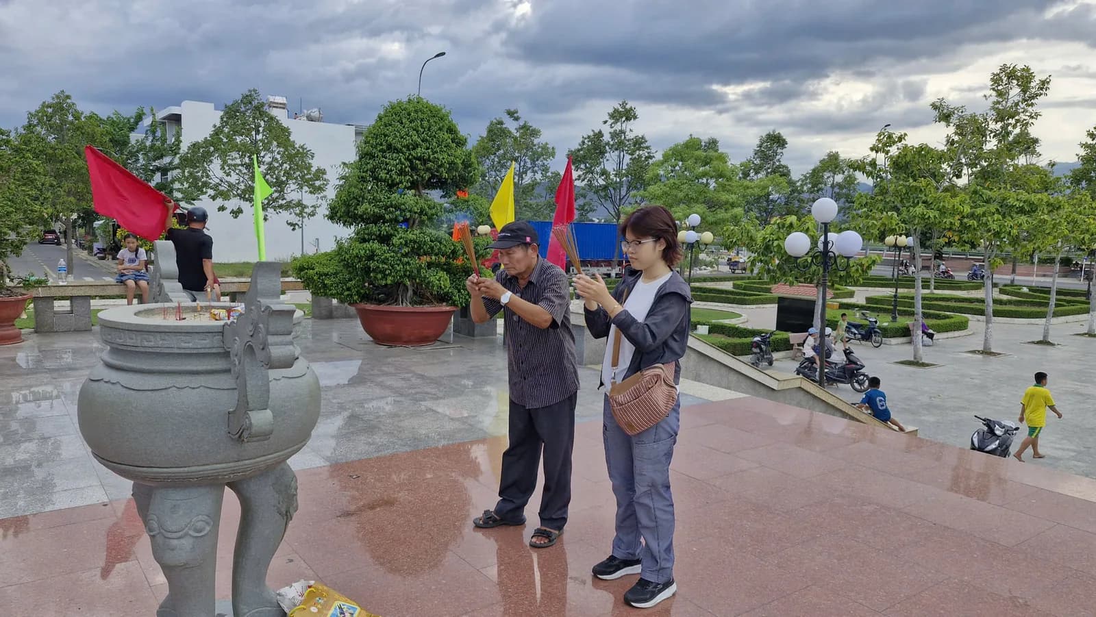 Một bác và một bạn trẻ cùng nâng bó hương trước lư hương lớn ở quảng trường.