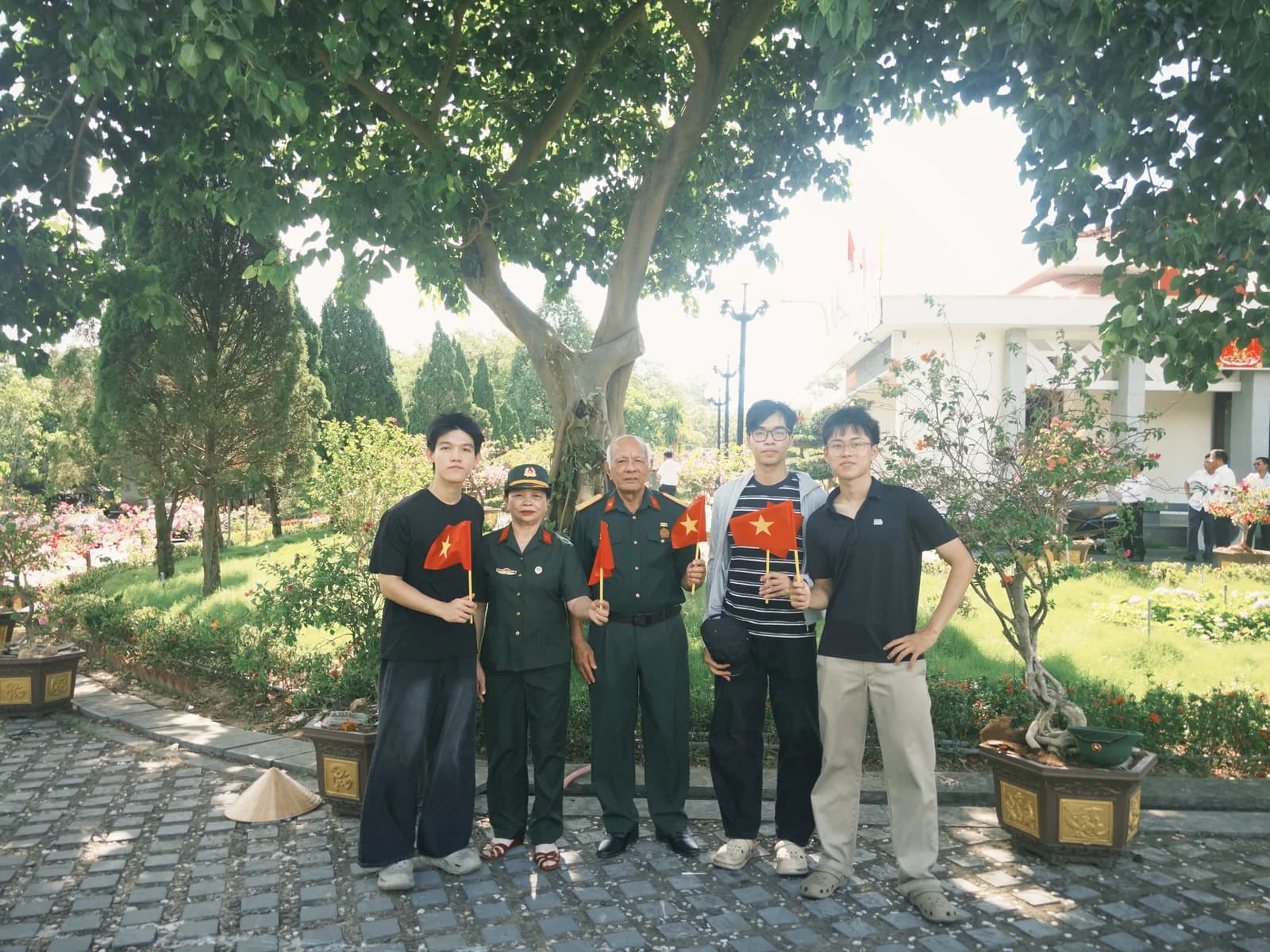 Các bạn trẻ và các bác cựu chiến binh đứng dưới tán cây trong khuôn viên Nghĩa trang Liệt sĩ Quốc gia Đường 9.