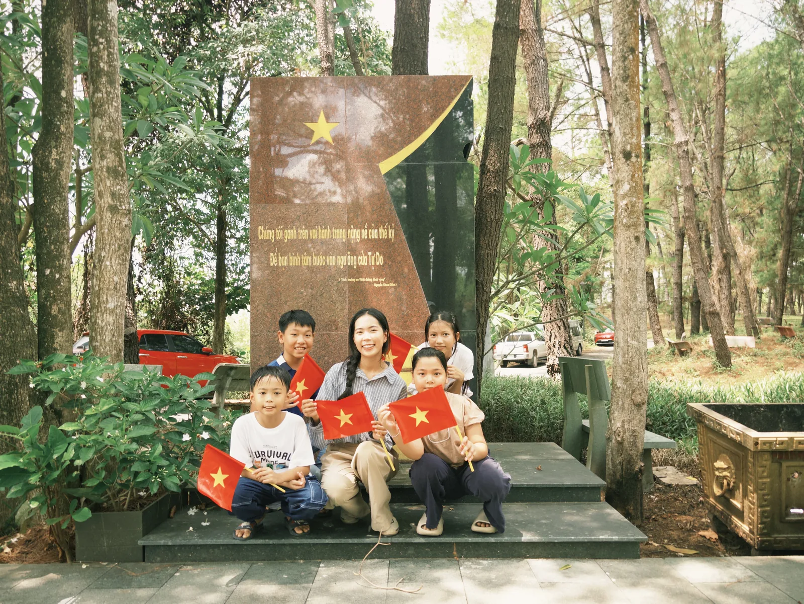 Một bạn trẻ và mấy em nhỏ cầm cờ Tổ quốc, ngồi bên tấm bia khắc thơ dưới rừng thông.