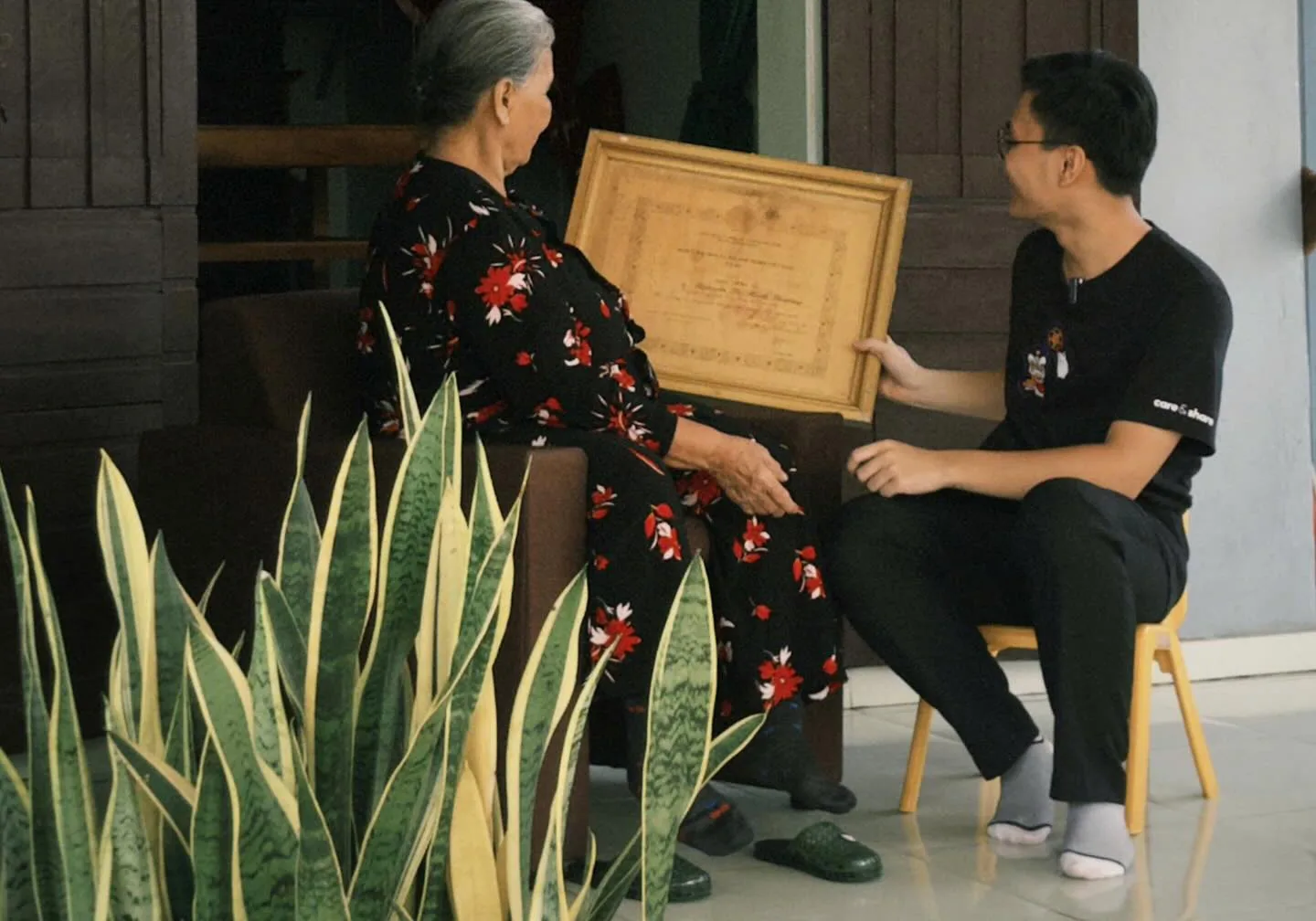 Một bạn trẻ ngồi cùng bà trong nhà, hai người cùng xem một tấm bằng đã cũ.