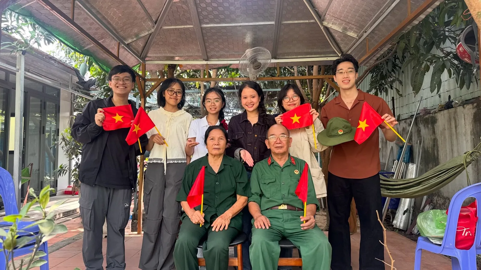 Sáu bạn trẻ đứng phía sau hai bác ngồi ghế, cả nhóm cùng cầm cờ Tổ quốc trong sân vườn.